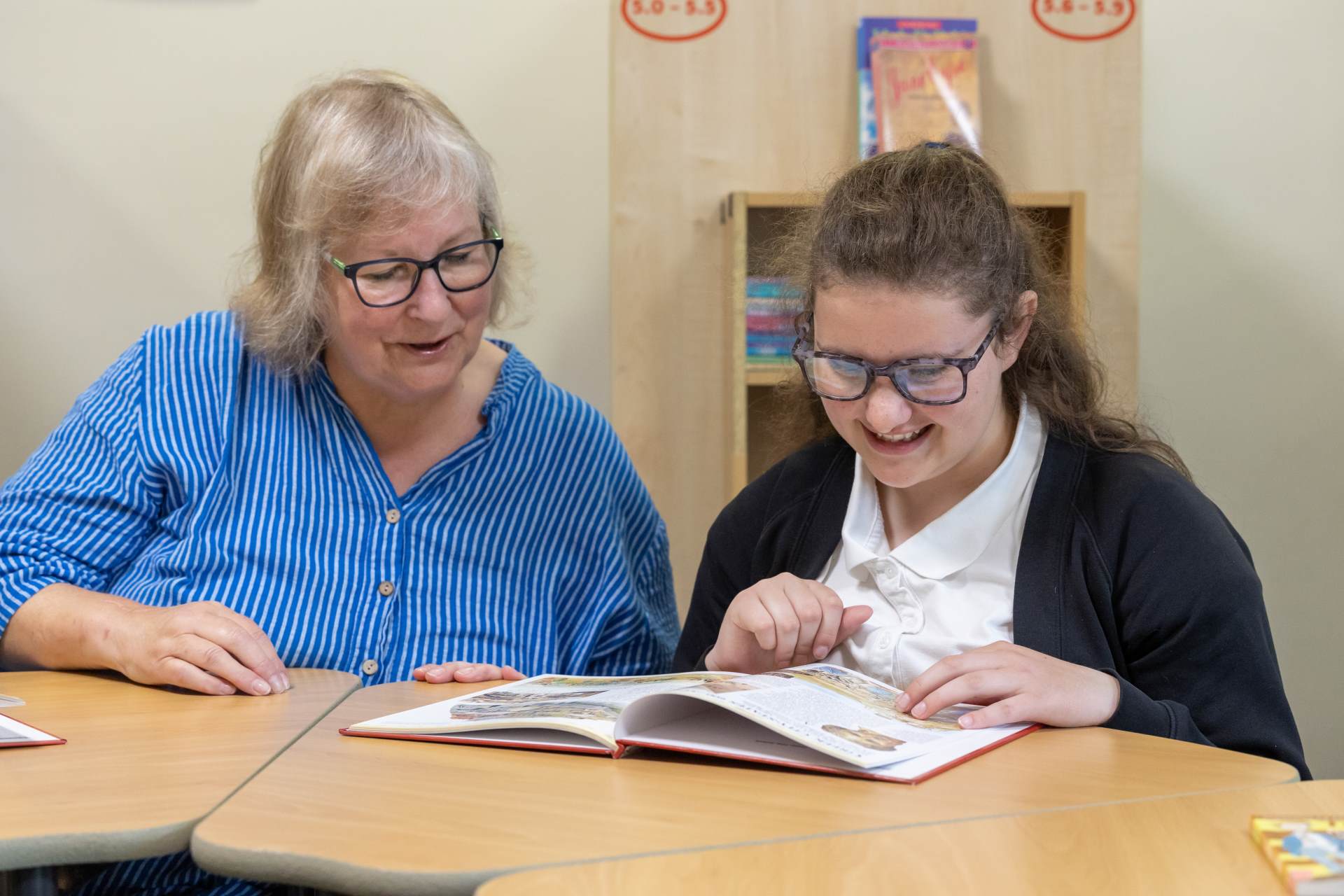 a woman with blonde/white hair and glasses and wearing a blue striped shirt is sitting next to a girl in a navy blue uniform. they are both looking at a book