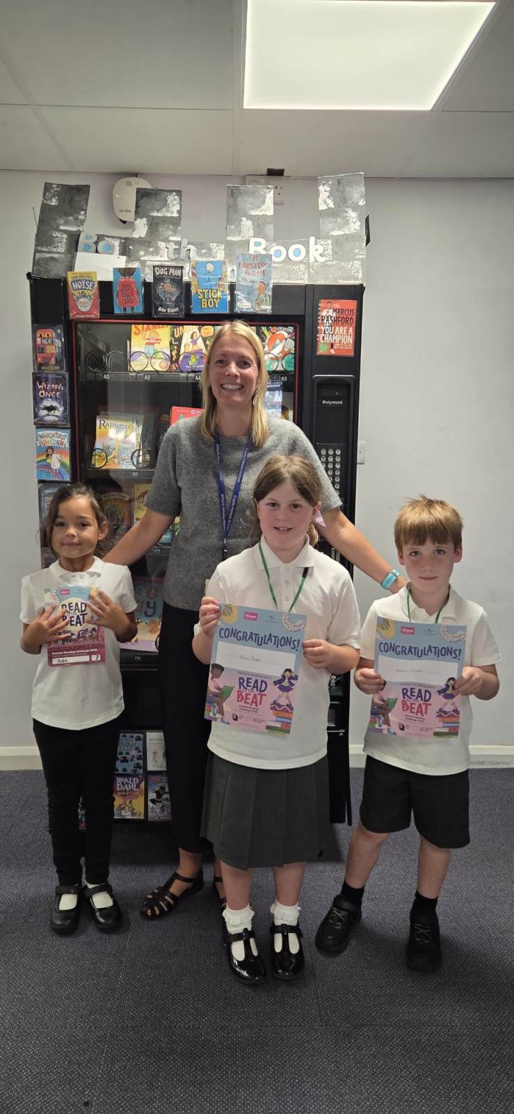 a woman with blonde hair and wearing a grey top is standing in front of a bookcase. she is smiling with three school pupils in front of her. They are all wearing white tops and grey skirts or trousers or leggings.