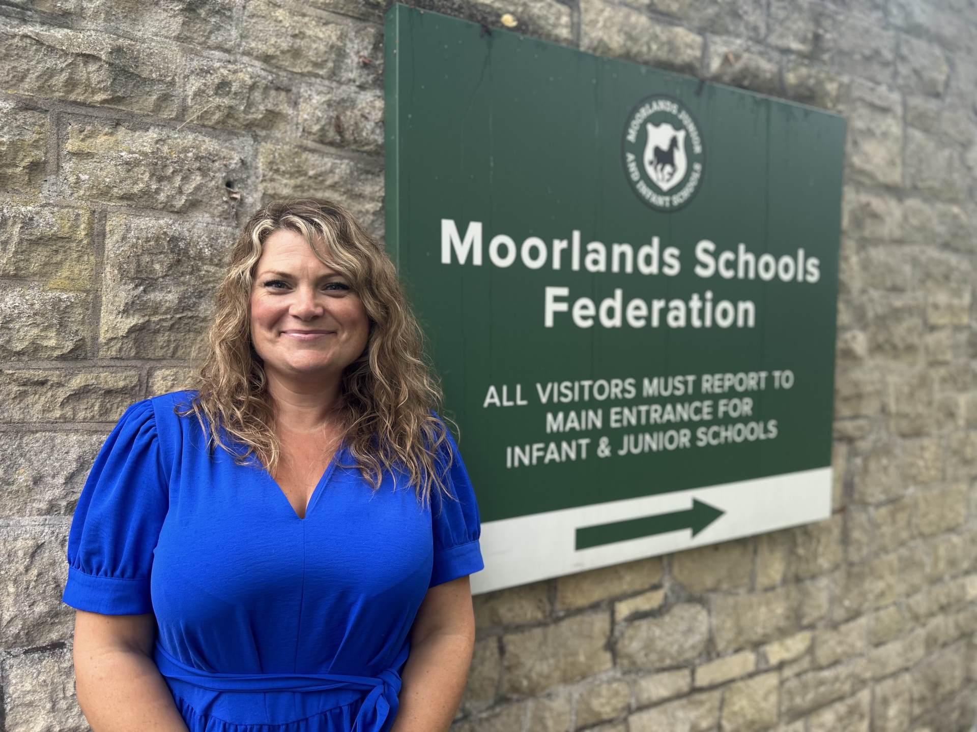Headteacher Moorlands School Bath a woman with blonde hair and wearing a blue dress stands beside a sign that says Moorlands Schools Federation